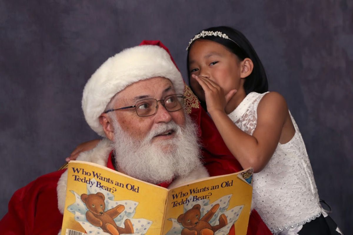 Santa Glenn reading a storybook while a young girl whispers in his ear