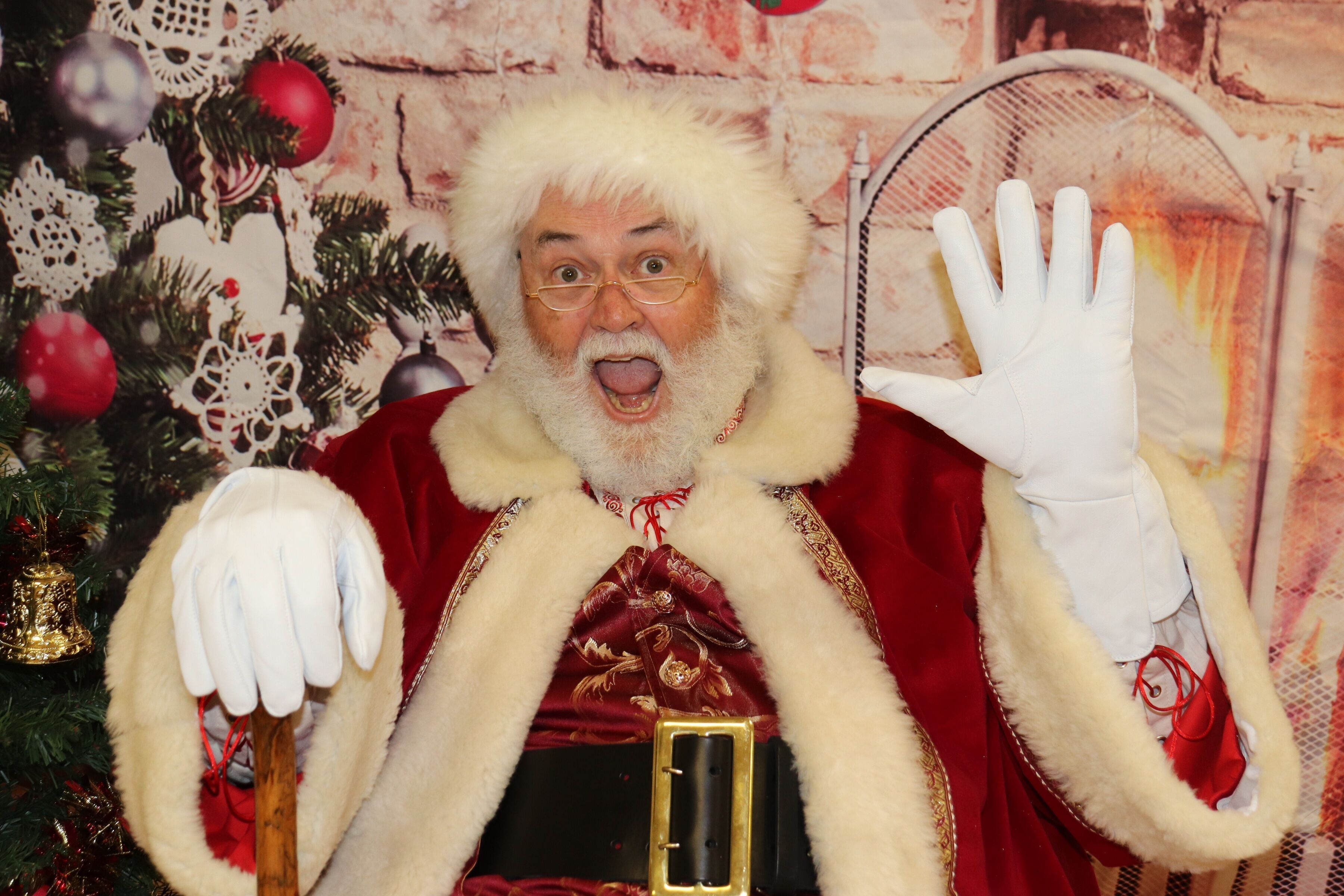 Santa Glenn waving with a white glove beside a decorated tree and fireplace
