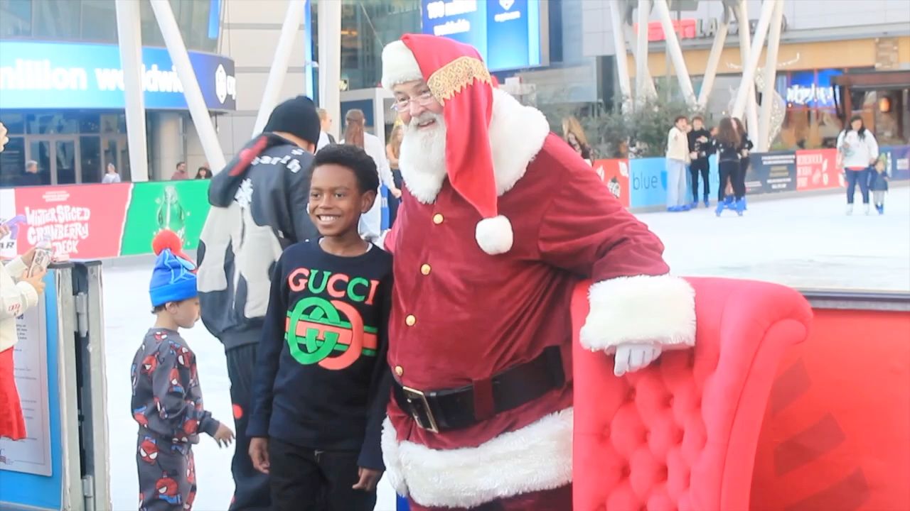 Santa Glenn smiling beside a beaming boy at an outdoor ice rink, next to his red velvet chair