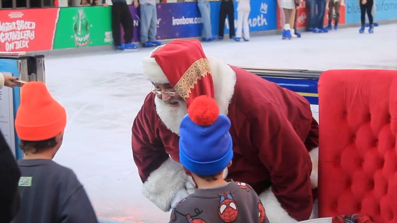 Santa Glenn kneeling to talk with a small boy in a blue knit hat at an ice rink